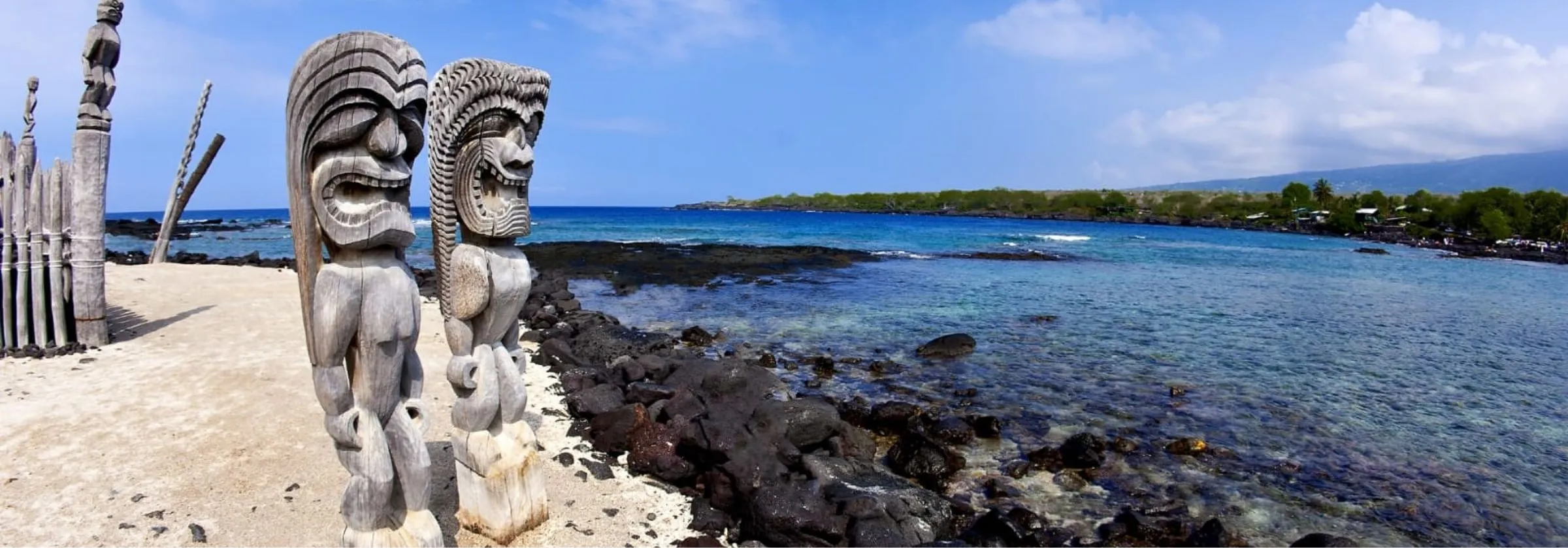 Hawaiian carved wooden statues (kiʻi) on a rocky beach by the ocean.