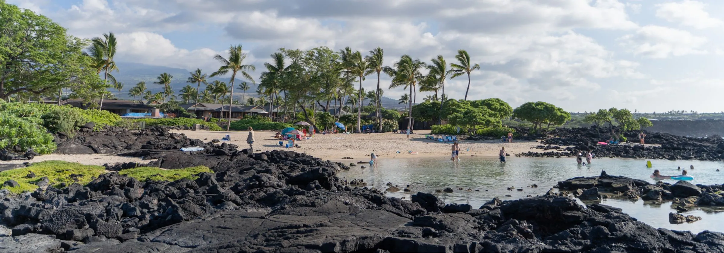 Sandy beach with palm trees, people swimming, and volcanic rocks in the foreground.