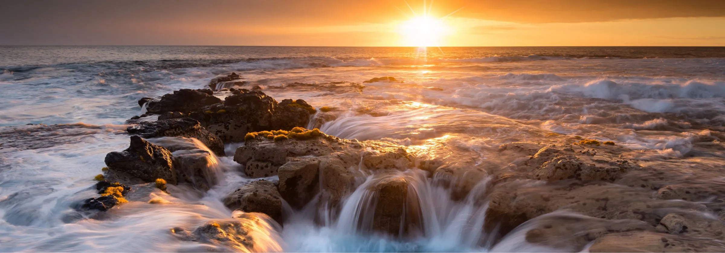 Sunset over ocean waves crashing on rocky shore.