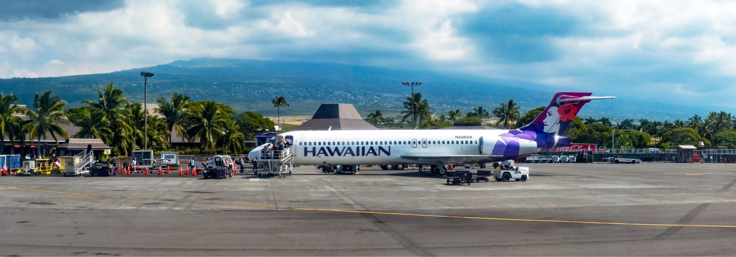 Hawaiian Airlines plane on a tarmac with palm trees and a mountain.