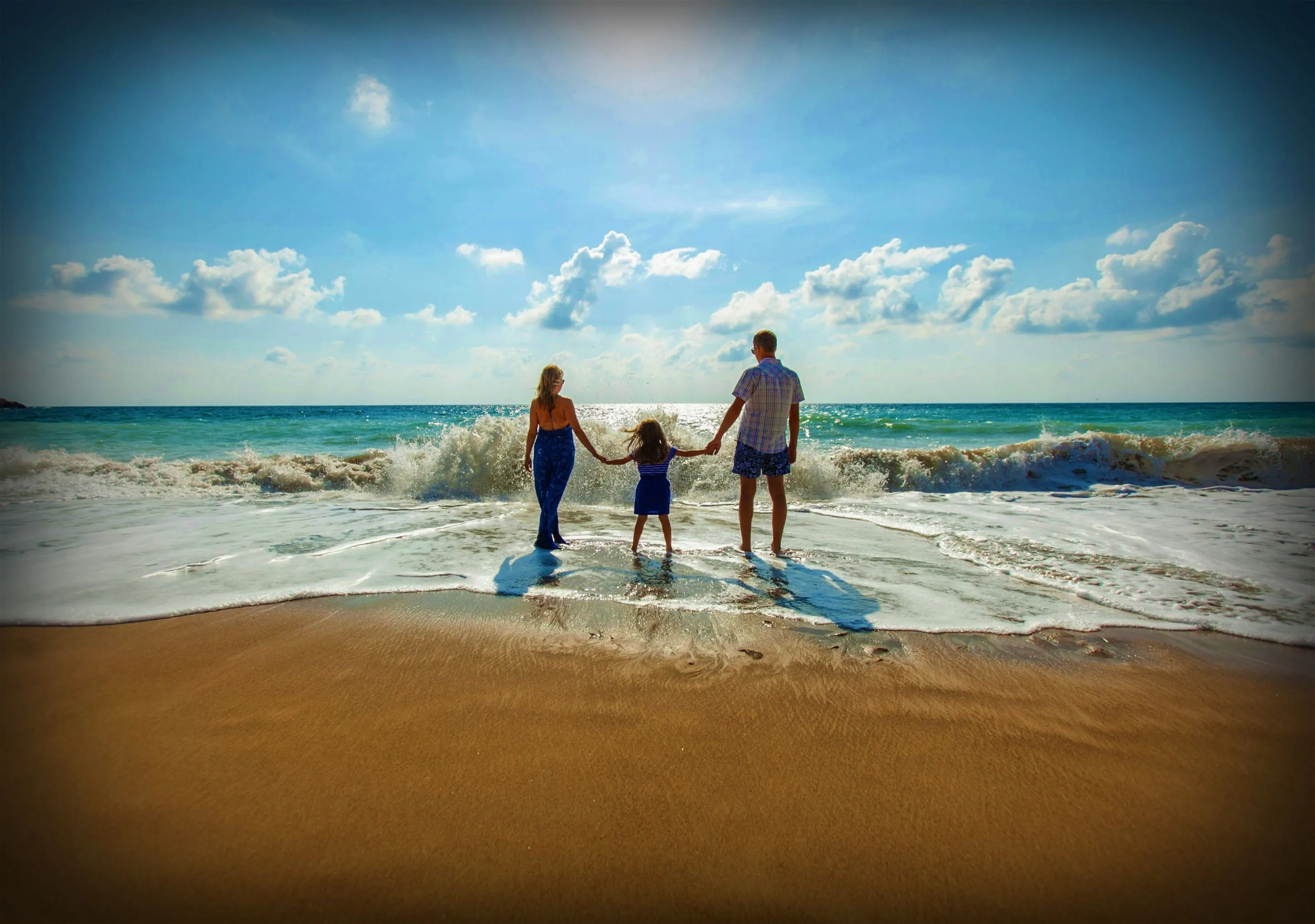 Family holding hands, standing on a beach, facing the ocean waves.
