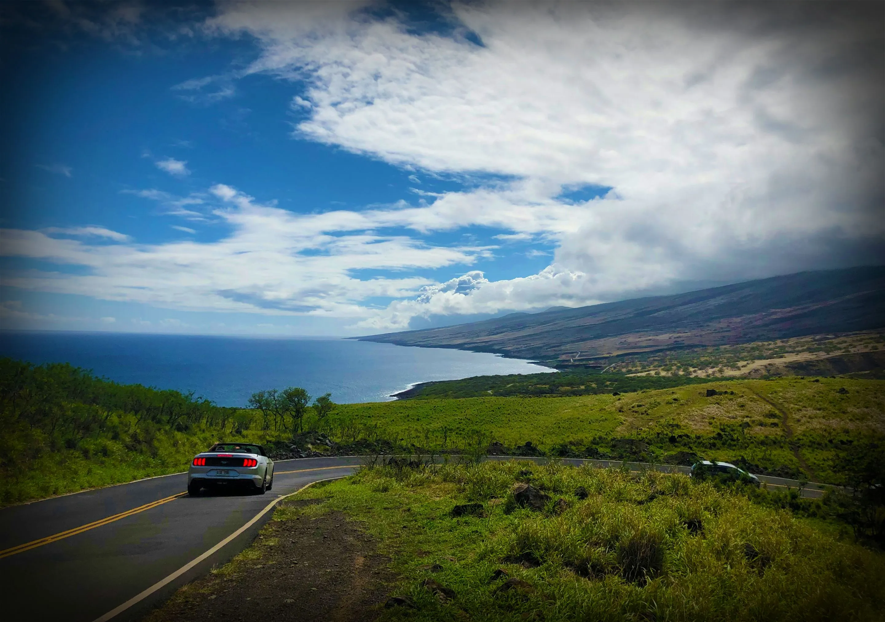 Car driving on a coastal road with ocean and mountain views.
