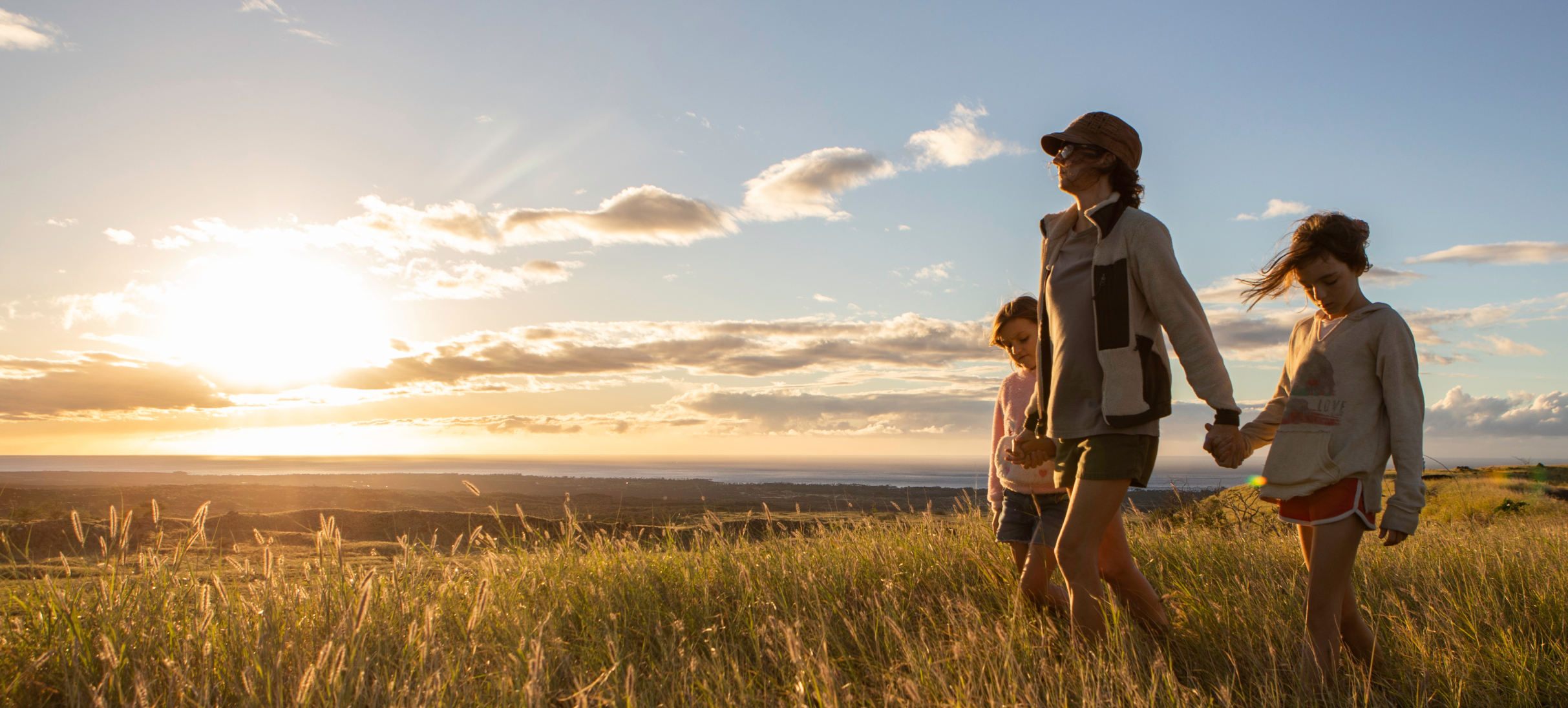 A person and two children walk hand in hand through a grassy field at sunset, with clouds painting the Hawaiian sky. This serene moment captures the essence of life near Nānā Kai, where Big Island new homes blend seamlessly with nature's beauty.