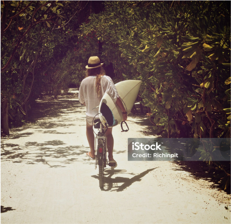 A person strolls along a sandy path, surrounded by lush greenery, walking a bike while balancing a surfboard. This scene captures the essence of Nānā Kai, where Hawaii new homes offer the perfect blend of adventure and tranquility on the Big Island.