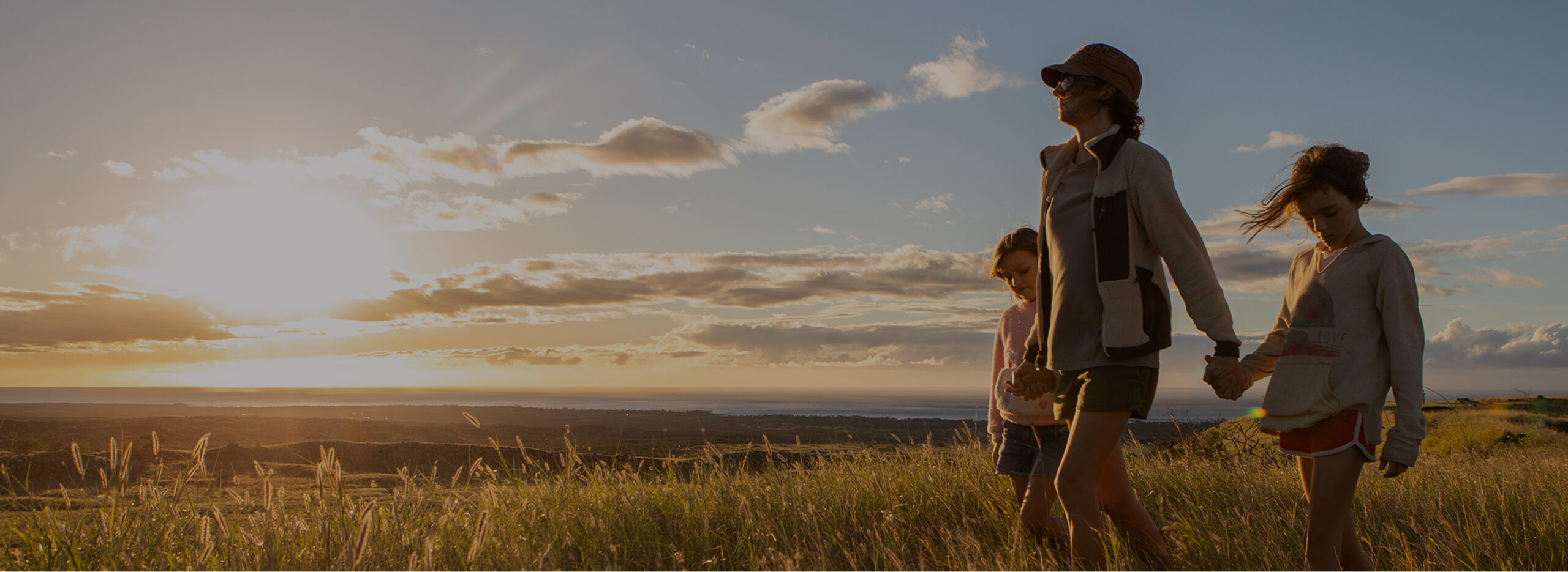 A person walks with two children through a grassy field at sunset under a partly cloudy sky.