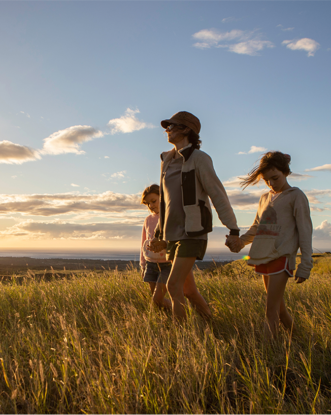 Three people walk through a grassy field at sunset, with one adult holding hands with two children.